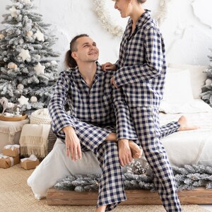 May include: Matching blue and white checkered pajama sets. The long-sleeved button-down shirts and pants are worn by a couple in a cozy bedroom setting. The room is decorated with Christmas trees and festive decor.