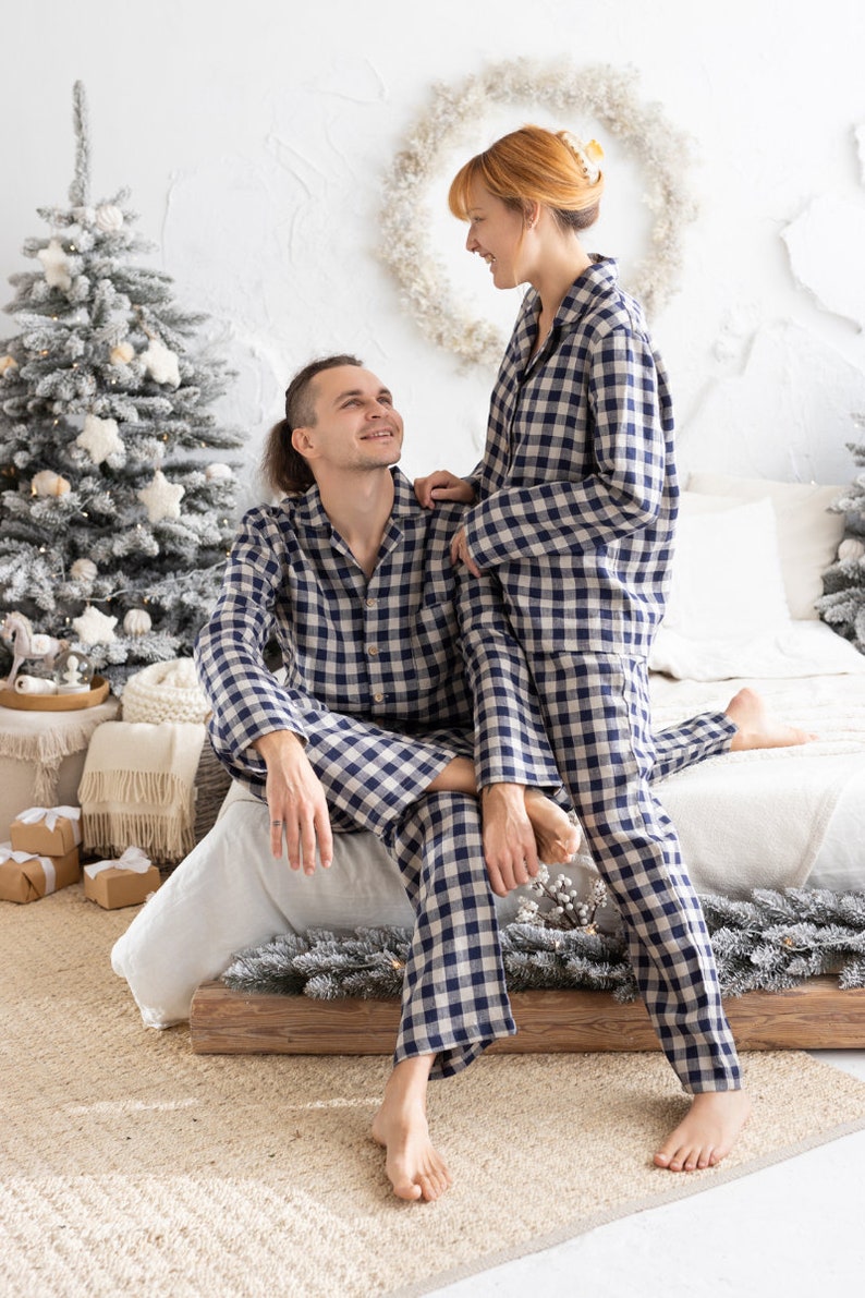 May include: Matching blue and white checkered pajama sets. The long-sleeved button-down shirts and pants are worn by a couple in a cozy bedroom setting. The room is decorated with Christmas trees and festive decor.