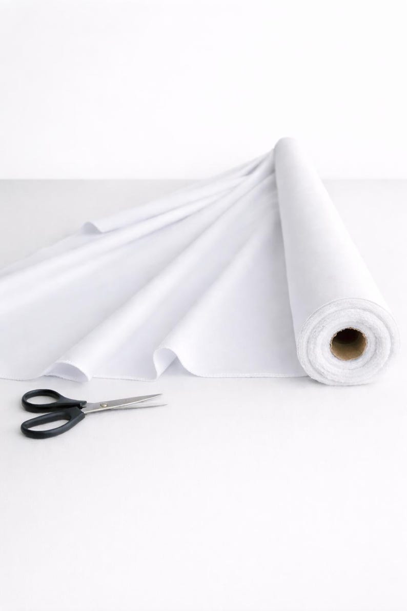 May include: A roll of solid white fabric partially unrolled to reveal a smooth, opaque texture. Beside the fabric roll lies a pair of black metal scissors with a pointed tip and ergonomic handles, ready for cutting the textile material.