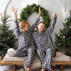 May include: A couple wearing matching navy and white checkered pajamas sit on a wooden bench, arms raised in celebration. A green wreath and Christmas trees decorate the background. A wrapped gift sits on the floor.