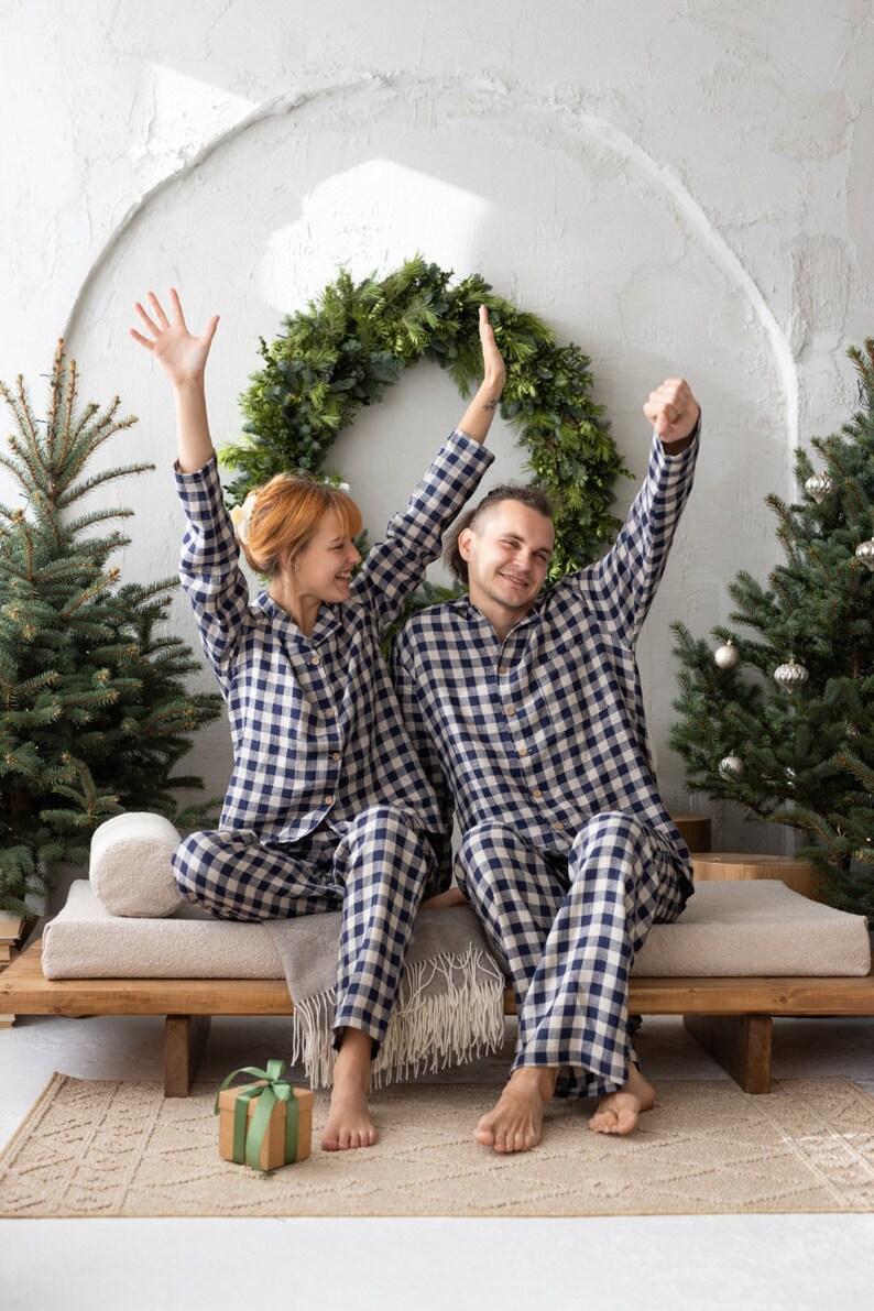 May include: A couple wearing matching navy and white checkered pajamas sit on a wooden bench, arms raised in celebration. A green wreath and Christmas trees decorate the background. A wrapped gift sits on the floor.