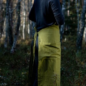 May include: A long, olive green linen apron with a large pocket in the front. The apron is tied at the waist and has a long strap that goes around the neck.
