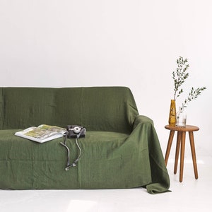 May include: A green linen sofa with a book and a camera on top. A small wooden stool with two glass vases and green branches sits to the right of the sofa.
