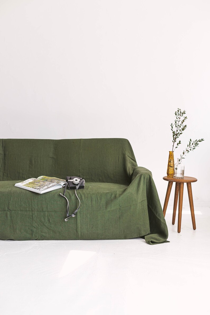 May include: A green linen sofa with a book and a camera on top. A small wooden stool with two glass vases and green branches sits to the right of the sofa.