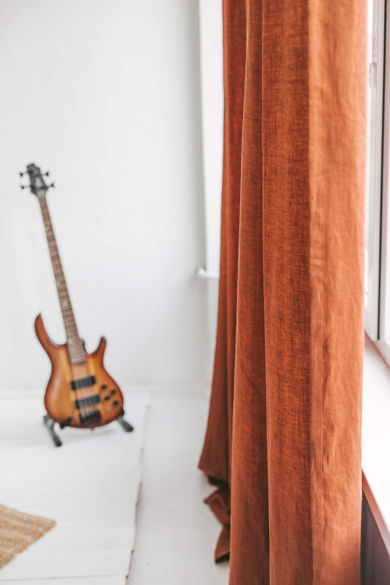 May include: A close-up of a rust-colored linen curtain hanging in a room with a blurred image of a bass guitar in the foreground.