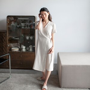 May include: A woman wearing a light beige linen wrap dress with short sleeves, standing in a room with a wooden cabinet and a light gray rug.