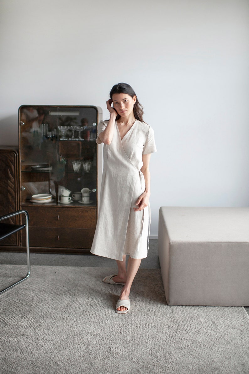 May include: A woman wearing a light beige linen wrap dress with short sleeves, standing in a room with a wooden cabinet and a light gray rug.