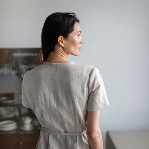 May include: A woman wearing a light beige linen dress with a tie belt and pockets. The dress has a simple, relaxed fit.