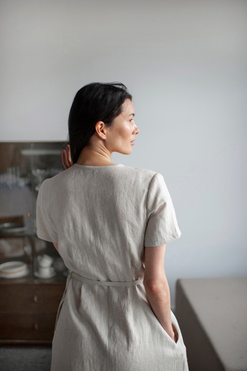 May include: A woman wearing a light beige linen dress with a tie belt and pockets. The dress has a simple, relaxed fit.
