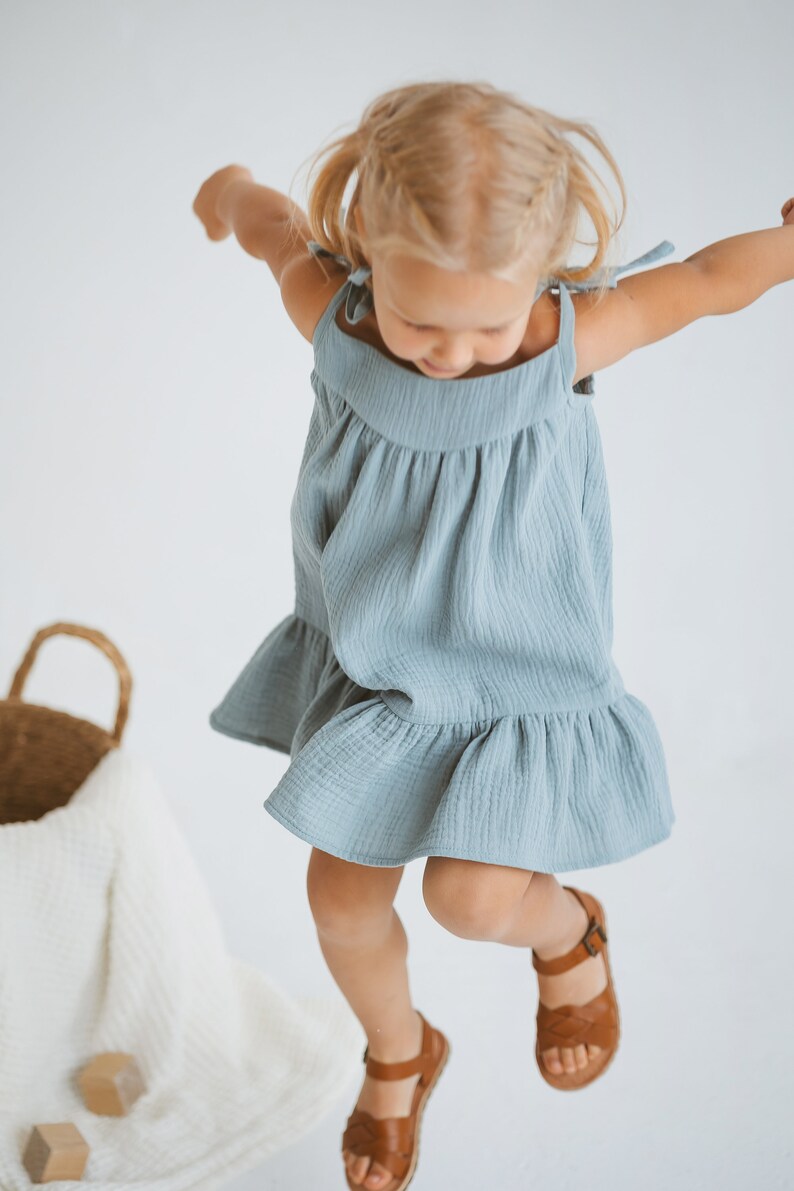 May include: A young child wearing a light blue dress with tie straps and a ruffled hem, paired with brown leather sandals. The child is jumping in the air. A woven basket and a white blanket are in the background.