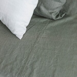 May include: A close-up of a green linen bed sheet with a white pillow on top. The sheet has a subtle texture and a seam running down the center.
