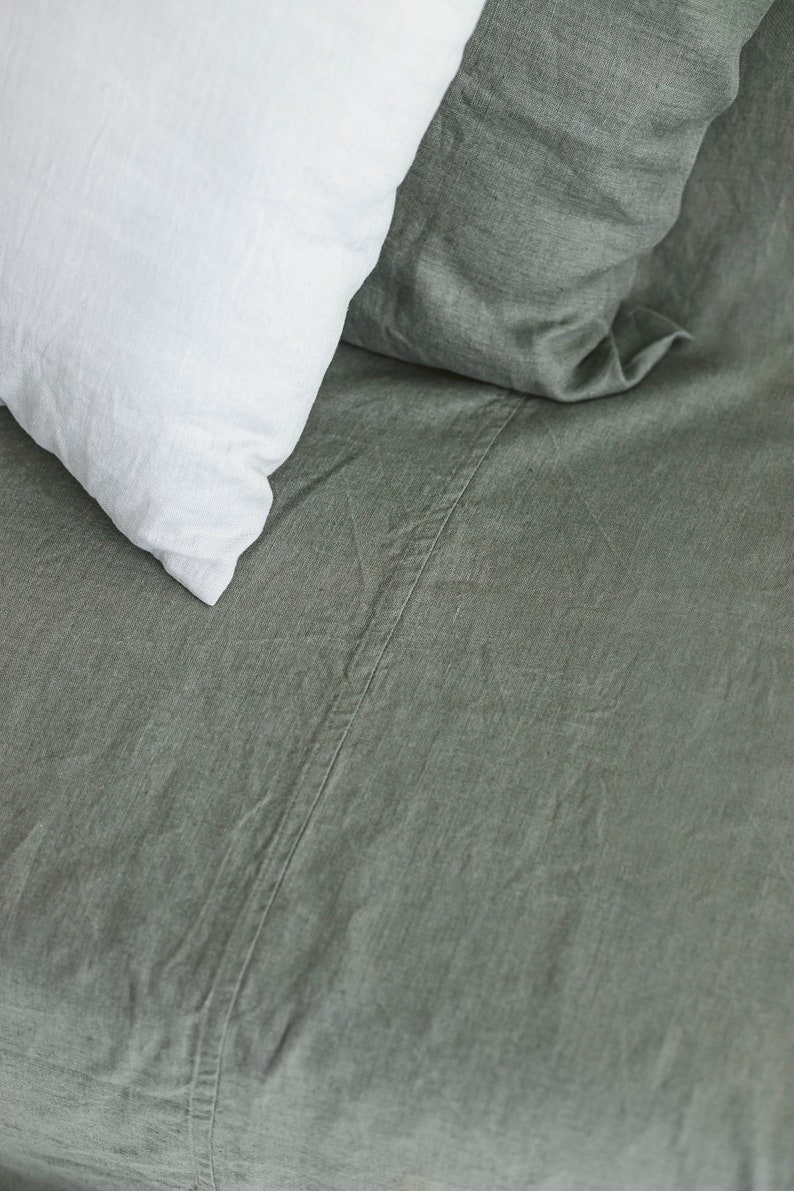 May include: A close-up of a green linen bed sheet with a white pillow on top. The sheet has a subtle texture and a seam running down the center.