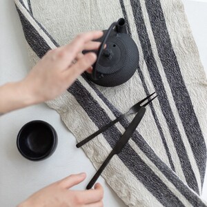 May include: A black cast iron teapot with a black and white striped linen tea towel, a black bowl, and two black metal utensils.
