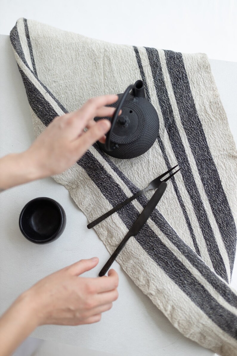 May include: A black cast iron teapot with a black and white striped linen tea towel, a black bowl, and two black metal utensils.
