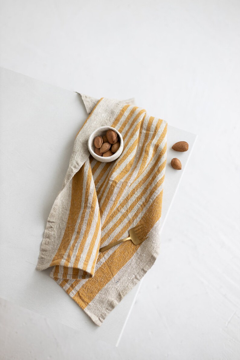May include: A yellow and white striped linen kitchen towel with a small white bowl of almonds and a gold fork on top.