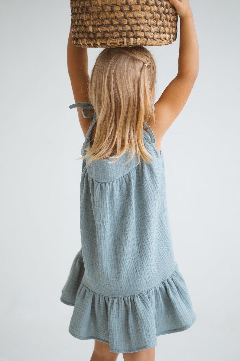 May include: A light blue dress with tie straps and a ruffled hem. The dress is made of a textured fabric and is being held up by a person. A woven basket is held above the head.