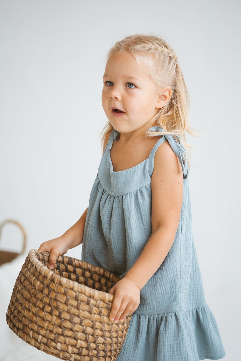 May include: A young child wearing a blue-green dress with tie straps holds a woven basket. The dress has a ruffled hem and a gathered bodice. The child has blonde hair styled in a braid and is looking upwards.