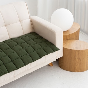 May include: A green quilted sofa cushion on a white sofa with a white lamp on a wooden table.