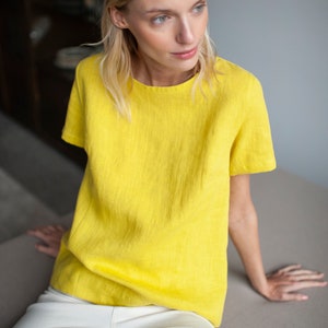 Può includere: Una donna indossa una blusa di lino giallo brillante a maniche corte e pantaloni bianchi. È seduta su un divano e guarda a destra.