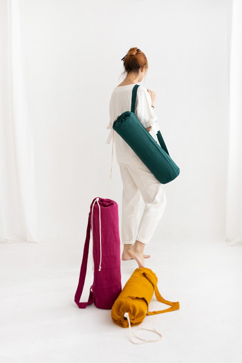 Puede incluir: Tres bolsas para esterillas de yoga en diferentes colores: verde, rosa y amarillo. Las bolsas est&aacute;n hechas de tela y tienen un cierre de cord&oacute;n. Las bolsas est&aacute;n sobre una superficie blanca.