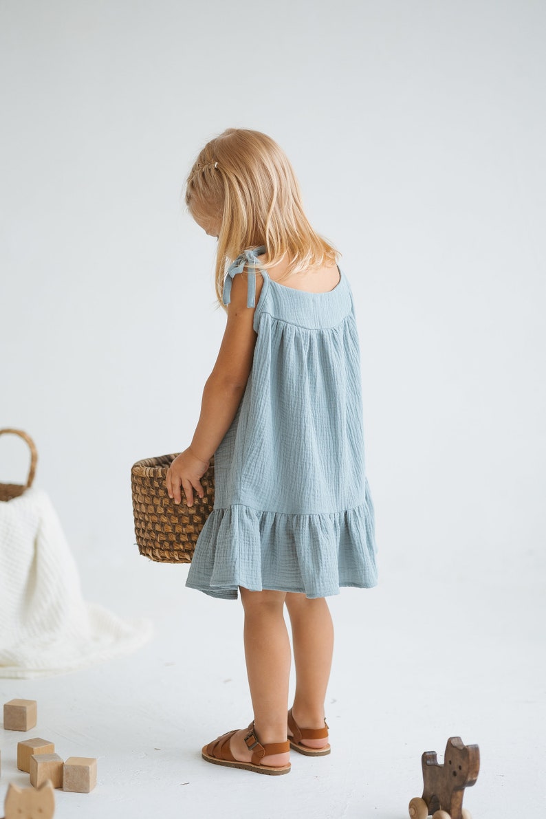May include: A light blue, ruffled hem dress with tie straps. The dress is worn with brown sandals. A woven basket is held in the hands. The dress is made of a textured fabric. The overall theme is children's clothing.