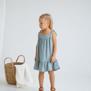 May include: A young child wearing a light blue, sleeveless dress with tie straps and a ruffled hem. The dress is paired with brown sandals. A woven basket with a white blanket and wooden toy dogs are in the background.
