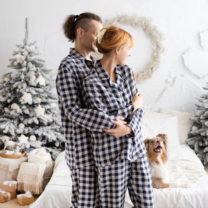 May include: A couple wearing matching navy and white checkered pajamas, embracing in a cozy bedroom setting. A Christmas tree and a dog are also in the frame. The pajamas are a classic sleepwear style.