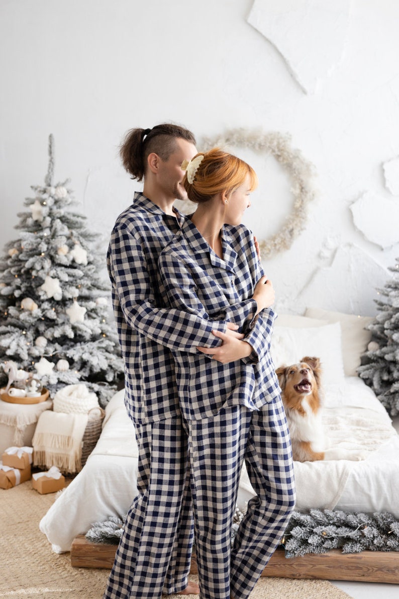 May include: A couple wearing matching navy and white checkered pajamas, embracing in a cozy bedroom setting. A Christmas tree and a dog are also in the frame. The pajamas are a classic sleepwear style.