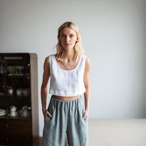 K&ouml;nnte beinhalten: Eine Frau tr&auml;gt ein wei&szlig;es Leinen-Crop-Top und eine Hose aus salbeigr&uuml;nem Leinen. Das Top hat einen Rundhalsausschnitt. Die Hose ist hoch tailliert, weit geschnitten und hat einen elastischen Bund. Das Outfit ist l&auml;ssig.