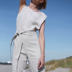 May include: A woman wearing a light beige linen jumpsuit with a wide belt. The jumpsuit has a relaxed fit and a sleeveless design. The woman is standing on a sandy beach with a blue sky in the background.