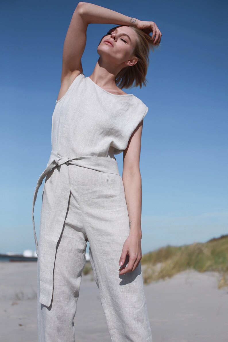May include: A woman wearing a light beige linen jumpsuit with a wide belt. The jumpsuit has a relaxed fit and a sleeveless design. The woman is standing on a sandy beach with a blue sky in the background.