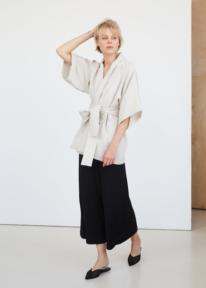 Może przedstawiać: Jasza w kolorze jasnobeżowym wykonana z lnu, z szerokim paskiem zawiązanym w talii. Kimono ma długie rękawy i luźny kr&oacute;j. Modelka ma na sobie czarne szerokie spodnie i czarne płaskie buty z siateczki.