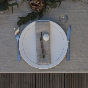 Pu&ograve; includere: Un piatto da cena in ceramica bianca con un tovagliolo di lino, un rametto di fiori secchi e un meringa bianca. Il piatto &egrave; posto su una tovaglia di lino grigia con una forchetta e un coltello d'argento.