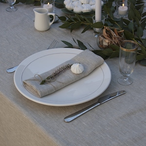 Pu&ograve; includere: Un piatto in ceramica bianca con un bordo dorato, un tovagliolo di lino, un piccolo rametto di fiori e un meringa bianca. Il piatto &egrave; posto su una tovaglia di lino grigio chiaro.
