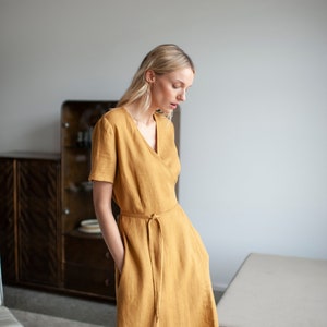 May include: A woman wearing a mustard yellow linen wrap dress with a V-neckline and short sleeves. The dress has a tie belt at the waist.