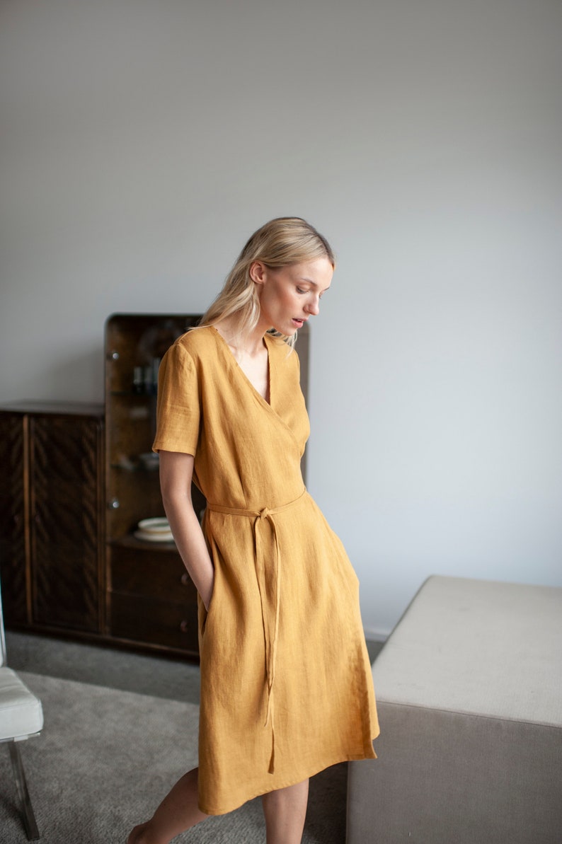 May include: A woman wearing a mustard yellow linen wrap dress with a V-neckline and short sleeves. The dress has a tie belt at the waist.