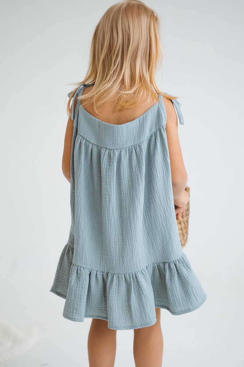 May include: A light blue dress with tie straps and a ruffled hem. The dress is made of a textured fabric and is worn by a young child. The child is facing away from the camera.