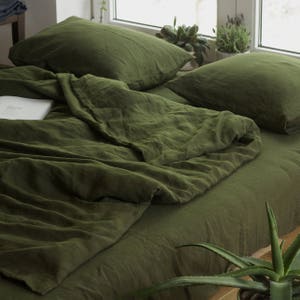 Op de afbeelding: Een groene linnen dekbedovertrek en twee bijpassende kussenslopen. De beddengoed is gerangschikt op een bed met een houten frame. Een potplant staat op de voorgrond.