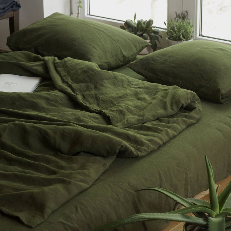 Op de afbeelding: Een groene linnen dekbedovertrek en twee bijpassende kussenslopen. De beddengoed is gerangschikt op een bed met een houten frame. Een potplant staat op de voorgrond.