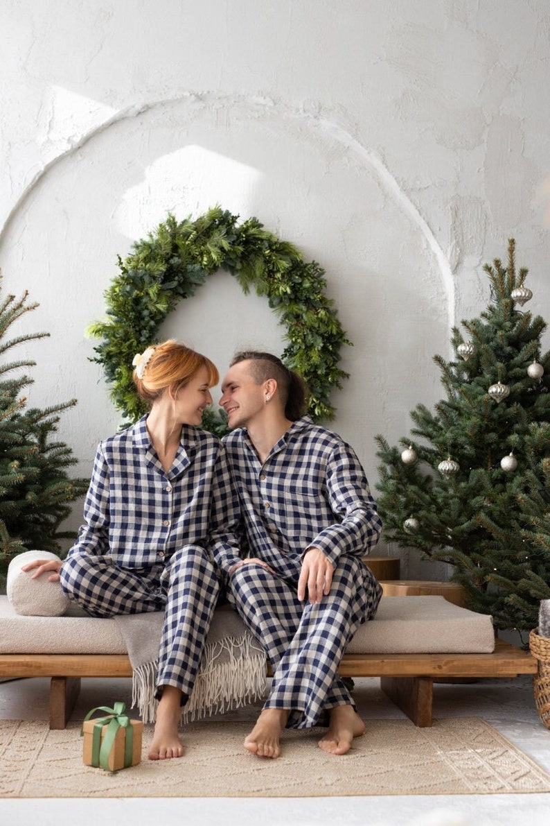 May include: A couple wearing matching navy and white plaid pajamas sit on a wooden bench. A green wreath hangs on the wall behind them, with a small Christmas tree to the right. A wrapped gift sits on the rug in front of them.
