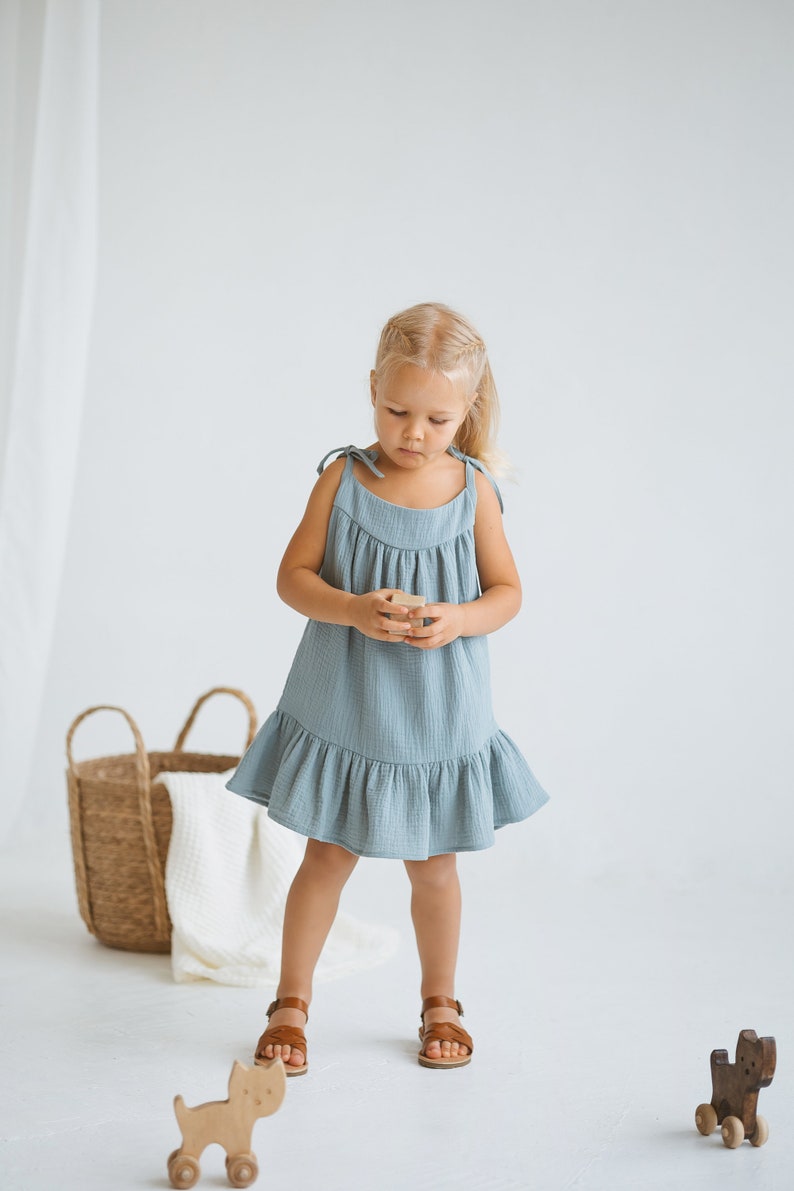 May include: A young child wearing a light blue dress with tie straps and a ruffled hem. The child is wearing brown sandals and holding a small wooden block. A woven basket and wooden toys are in the background.