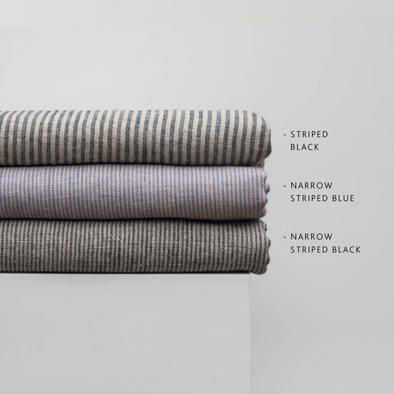 May include: Three rolls of fabric with vertical stripes in black, blue, and black. The top roll is striped black, the middle roll is narrow striped blue, and the bottom roll is narrow striped black. The fabric is stacked on a white surface.