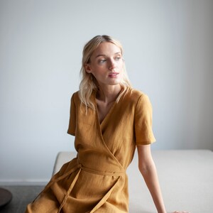 May include: A woman wearing a mustard yellow linen wrap dress, sitting on a light gray couch. She is looking to the right of the frame.