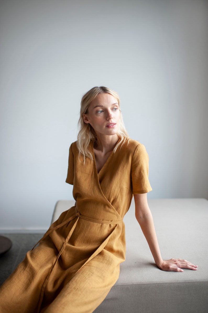 May include: A woman wearing a mustard yellow linen wrap dress, sitting on a light gray couch. She is looking to the right of the frame.