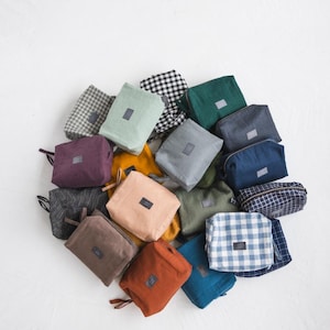May include: A collection of small, square fabric pouches in various colors and patterns, including solid hues like teal, mustard, and rust, and checkered and striped designs. Each pouch has a small tag and a zipper or drawstring closure.