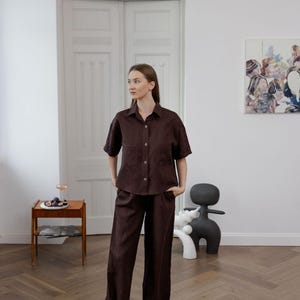 K&ouml;nnte beinhalten: Ein dunkelbraunes Leinenhemd und eine passende Hose mit weitem Bein. Das kurz&auml;rmelige Hemd hat zwei Vordertaschen und Knopfverschl&uuml;sse. Das Outfit wird in einem hellen Raum mit Holzboden und einem modernen Kunstwerk an der Wand getragen.