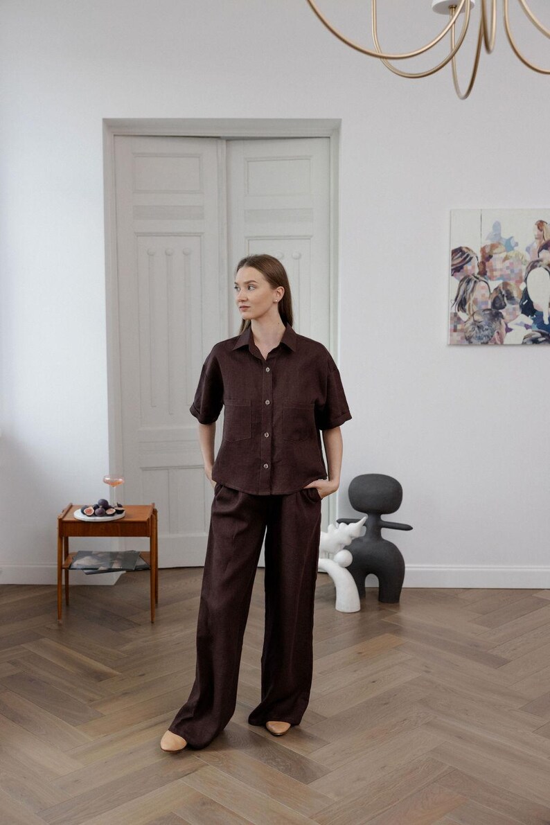 K&ouml;nnte beinhalten: Ein dunkelbraunes Leinenhemd und eine passende Hose mit weitem Bein. Das kurz&auml;rmelige Hemd hat zwei Vordertaschen und Knopfverschl&uuml;sse. Das Outfit wird in einem hellen Raum mit Holzboden und einem modernen Kunstwerk an der Wand getragen.