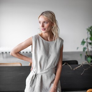 May include: A woman wearing a light beige linen jumpsuit with a tie belt. The jumpsuit has a sleeveless design and a relaxed fit. The woman is standing in front of a black table.
