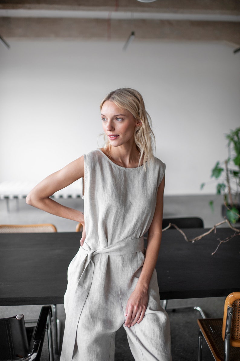 May include: A woman wearing a light beige linen jumpsuit with a tie belt. The jumpsuit has a sleeveless design and a relaxed fit. The woman is standing in front of a black table.