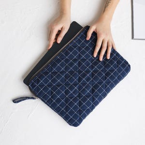 Puede incluir: Una funda para portátil acolchada de color azul marino con cierre de cremallera y un patrón de cuadrícula blanca. La funda está siendo sostenida por dos manos.